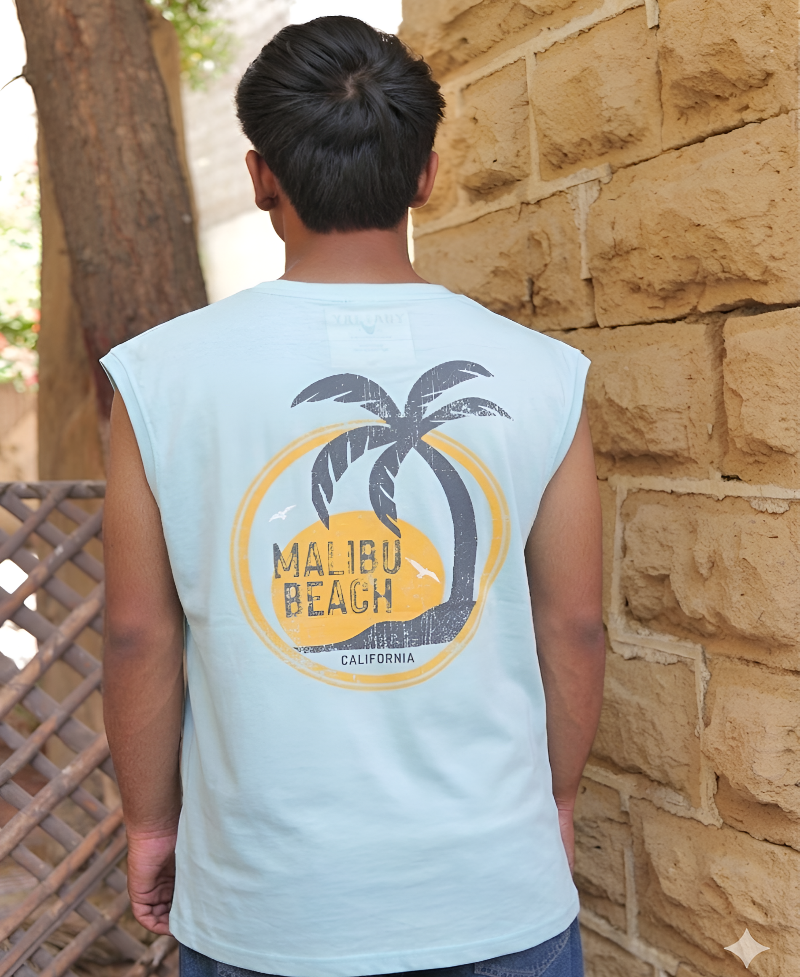 Person wearing a sleeveless shirt with a palm tree and 'Malibu Beach, California' design, standing against a stone wall.