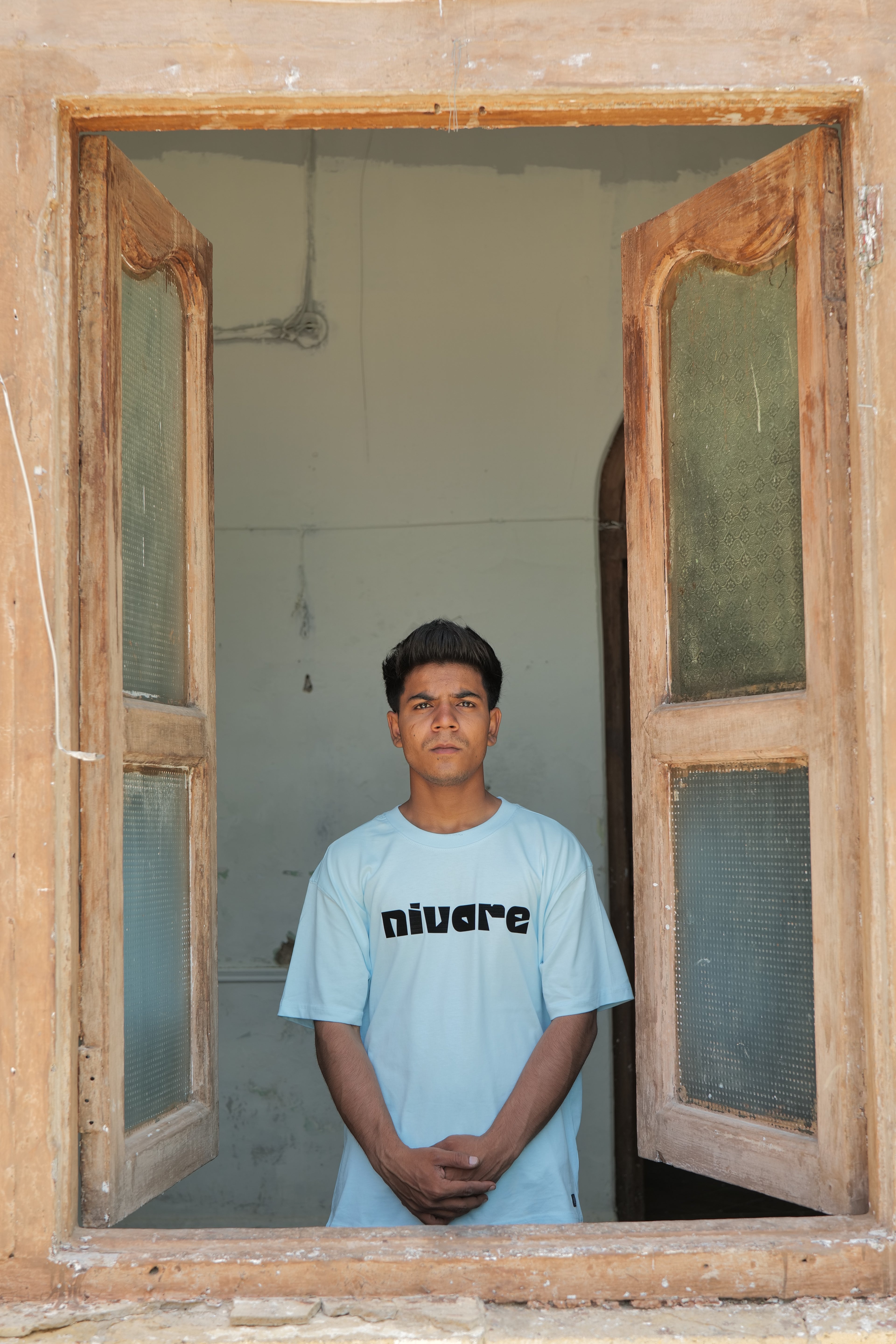 Person wearing a light blue t-shirt with text standing inside a room through an open wooden door.