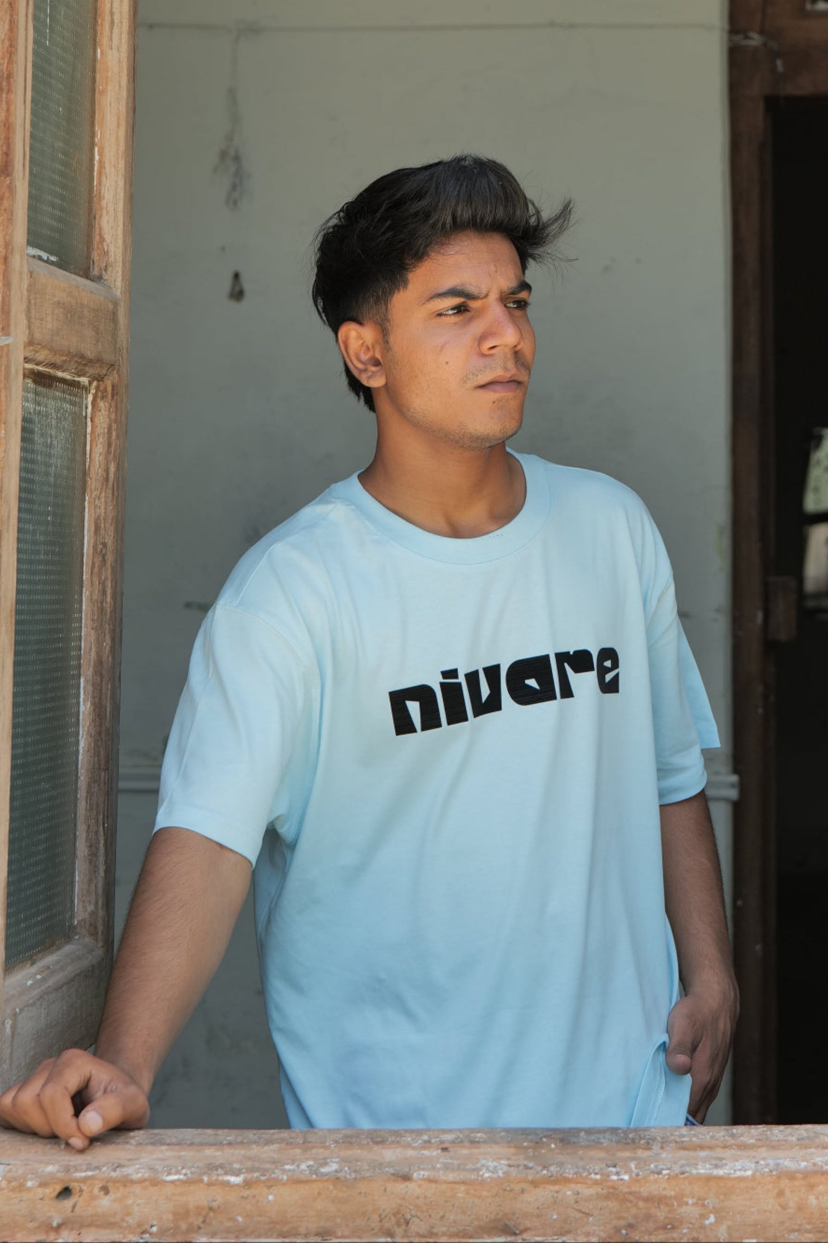 Person wearing a light blue t-shirt with 'nivare' printed on it, standing in a doorway.