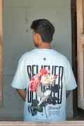 Person wearing a light blue t-shirt with floral design and black text, standing inside a wooden-framed room.