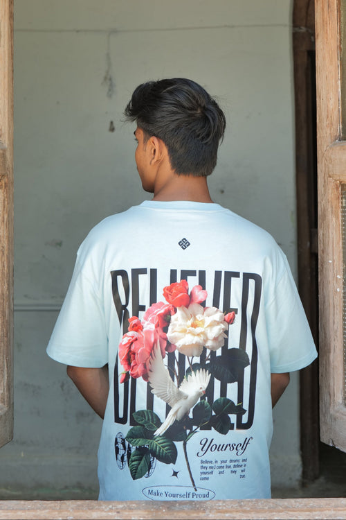 Person wearing a light blue t-shirt with floral design and black text, standing inside a wooden-framed room.