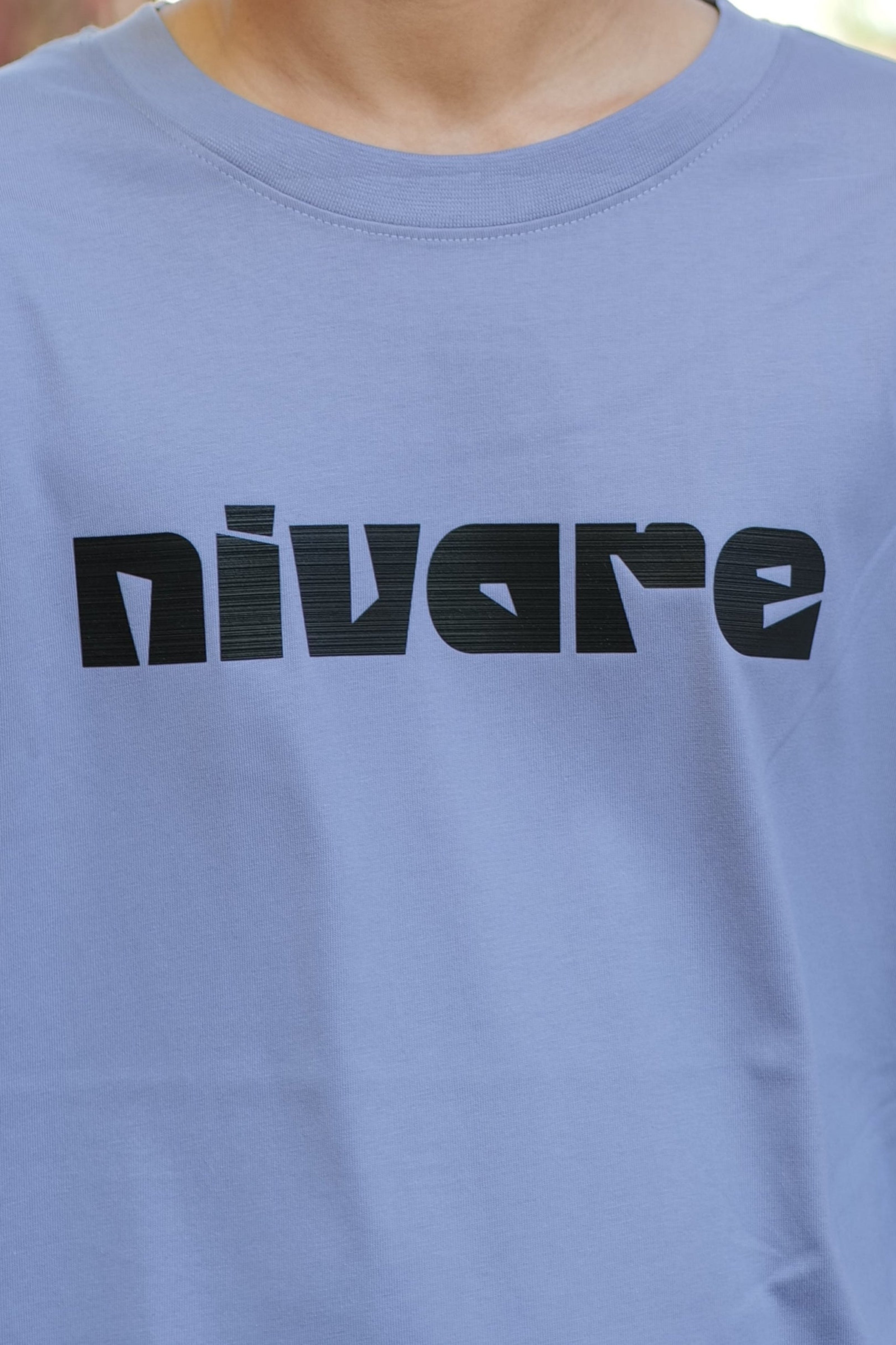 Person wearing a light blue t-shirt with 'Divare' logo, standing outdoors near stone wall and tree.