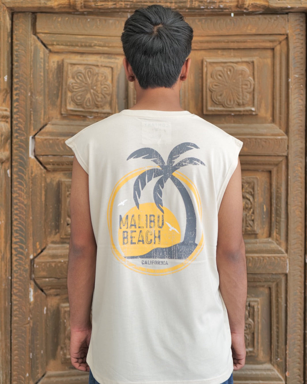 Person wearing a sleeveless shirt with 'Malibu Beach' design in front of a wooden door.