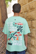 Person wearing a light green t-shirt with a colorful design and text, standing against a stone wall.
