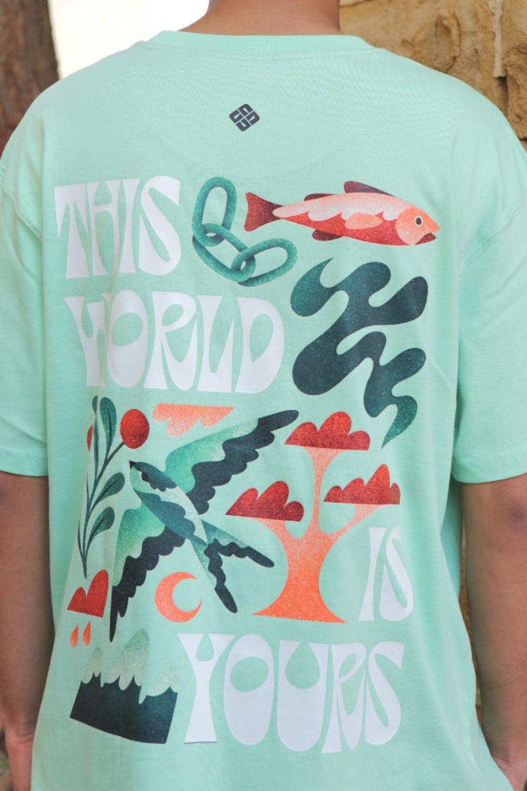 Person wearing a light green t-shirt with a colorful design and text, standing against a stone wall.