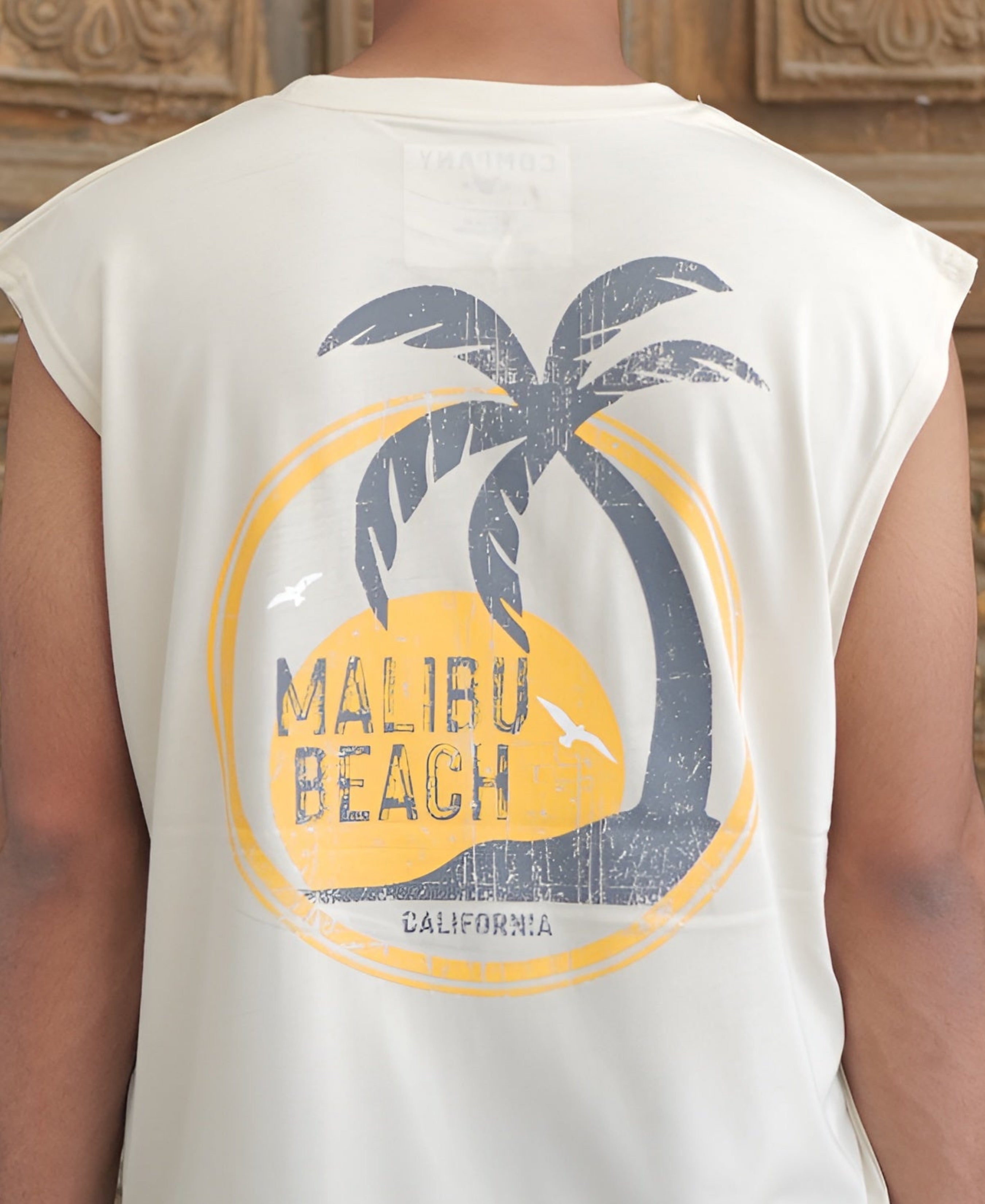 Person wearing a white sleeveless shirt with 'Malibu Beach' design in front of a wooden door.