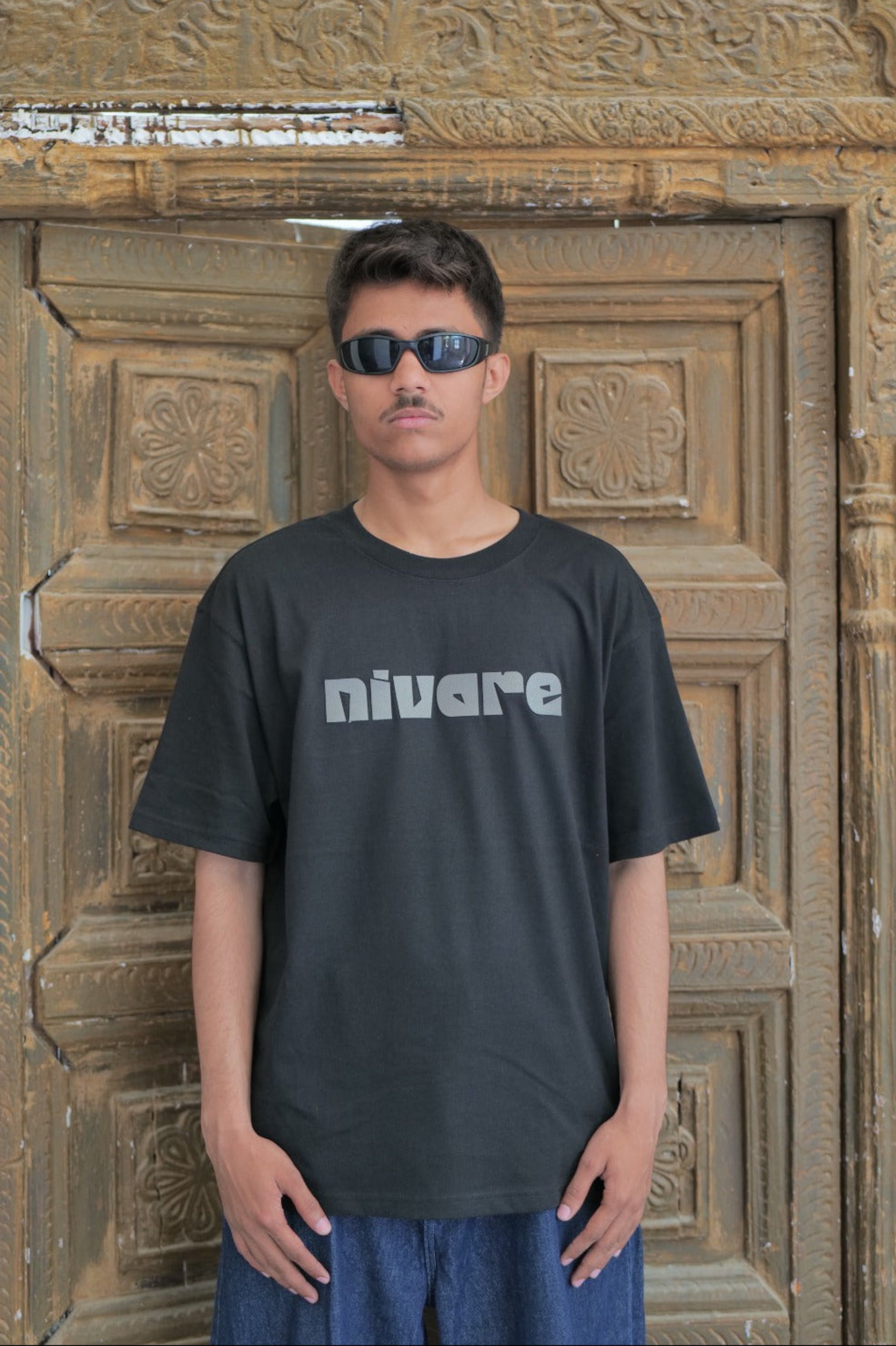 Person wearing a black t-shirt with 'nivare' logo in front of an ornate wooden door.