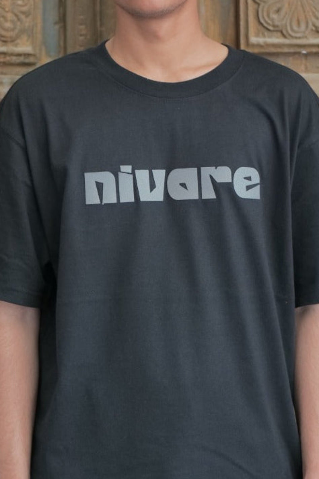 Person wearing a black t-shirt with 'nivare' logo standing in front of an ornate wooden door.