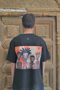 Person wearing a black t-shirt with a colorful graphic design of three people on the back, standing in front of an ornate wooden door.