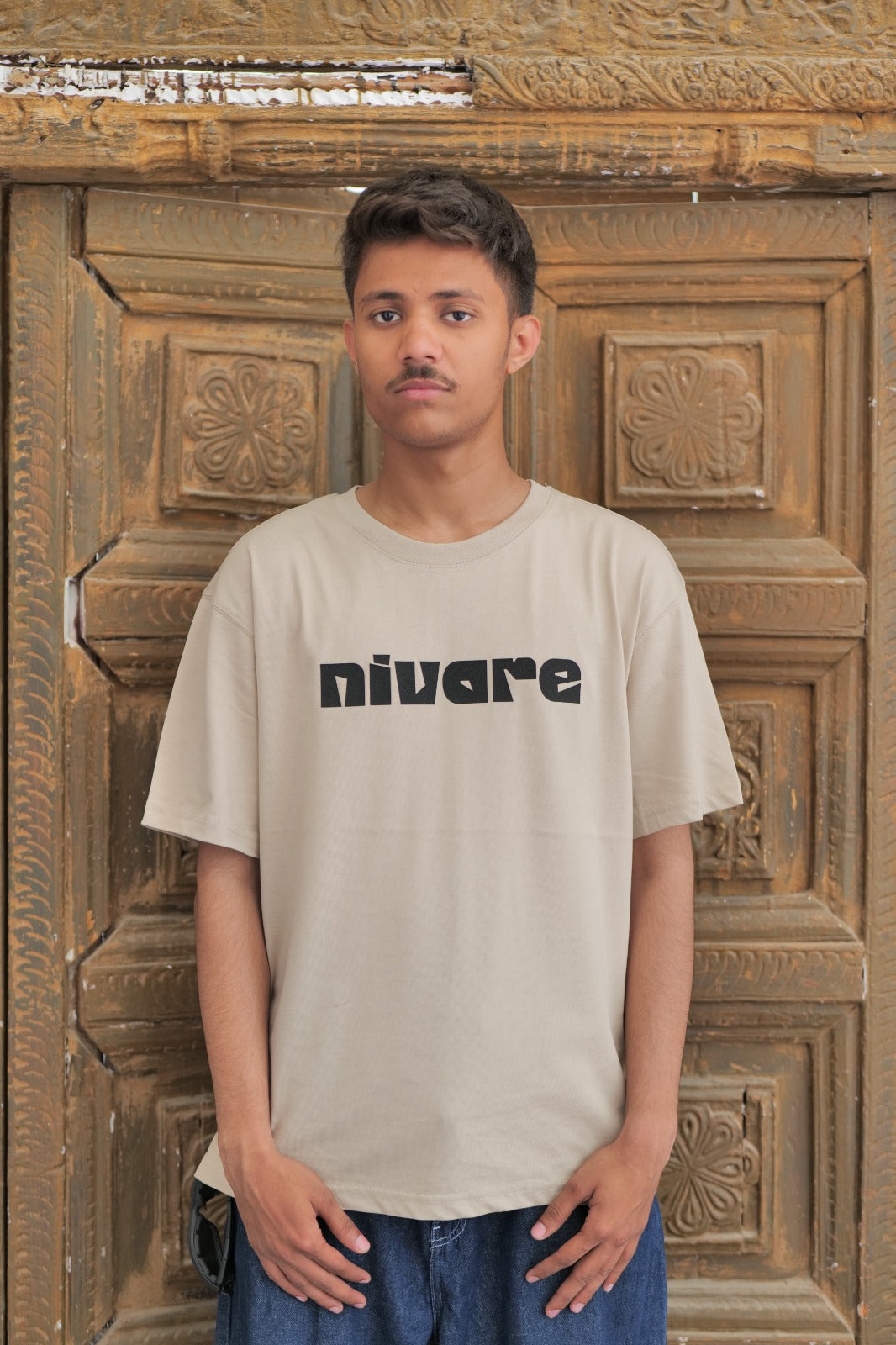 Person wearing a beige t-shirt with 'nivare' logo in front of a wooden door.