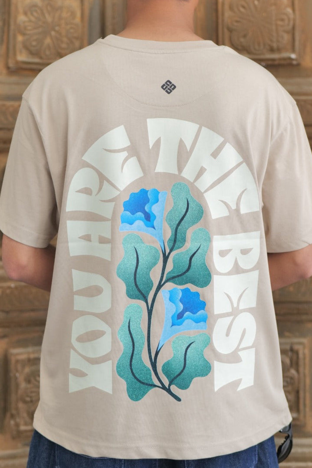 Person wearing a beige t-shirt with a floral design on the back, standing in front of a wooden door.