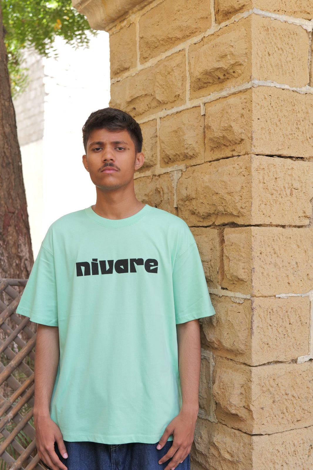 Person wearing a light green t-shirt with 'nivare' printed on it, standing against a stone wall.