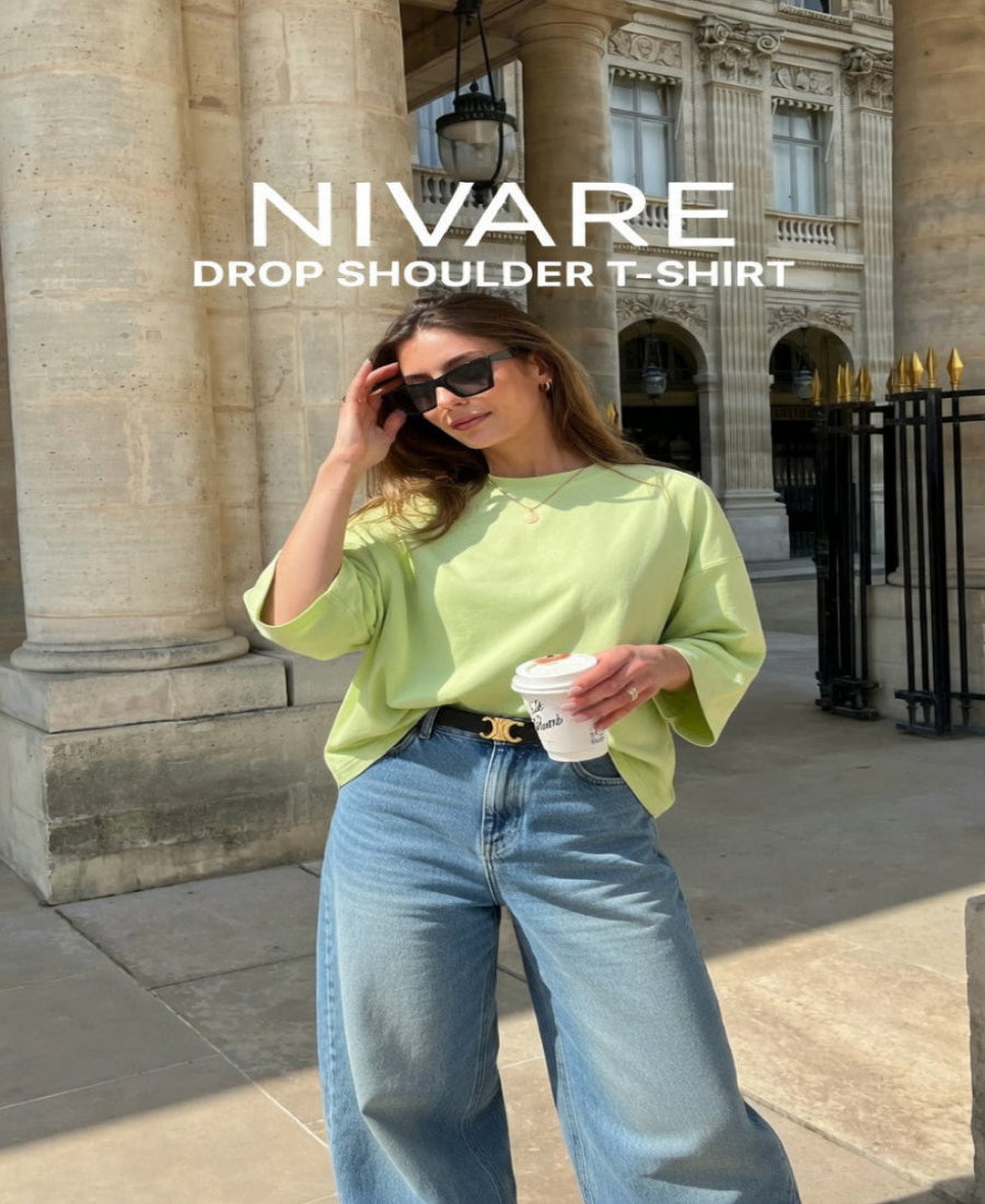 Woman wearing a green drop shoulder t-shirt and blue jeans, standing in front of classical architecture.