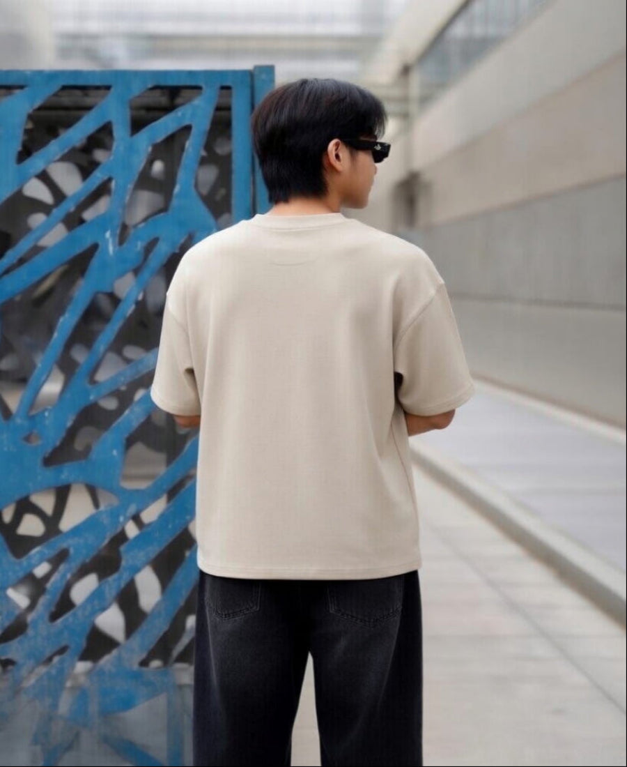 Person wearing a beige t-shirt and sunglasses, standing in front of a textured wall.