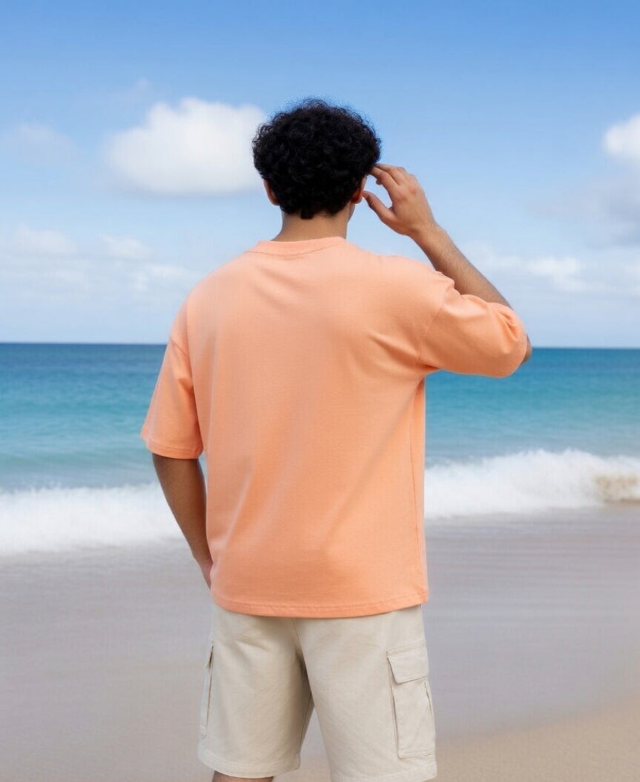 Person wearing an orange shirt and beige shorts standing on a beach with clear blue sky and ocean.