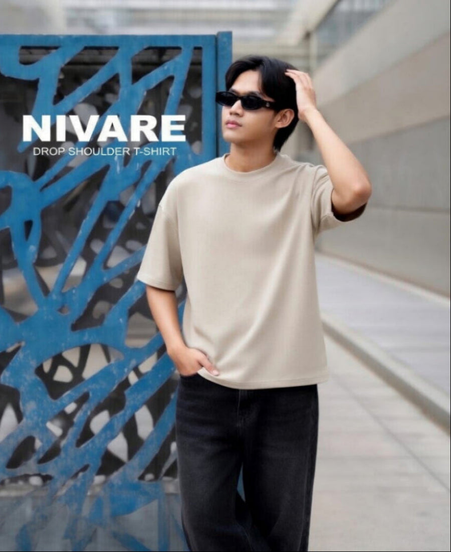 Person wearing a beige drop shoulder t-shirt with 'NIVARE' branding in an urban setting.