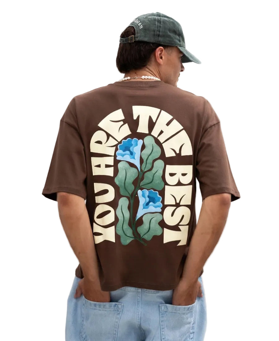Person wearing a brown t-shirt with a floral design and text on the back, standing against a plain background.