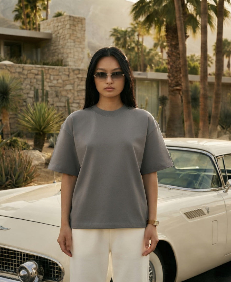 Person wearing a gray t-shirt and sunglasses standing in front of a vintage car with palm trees in the background.