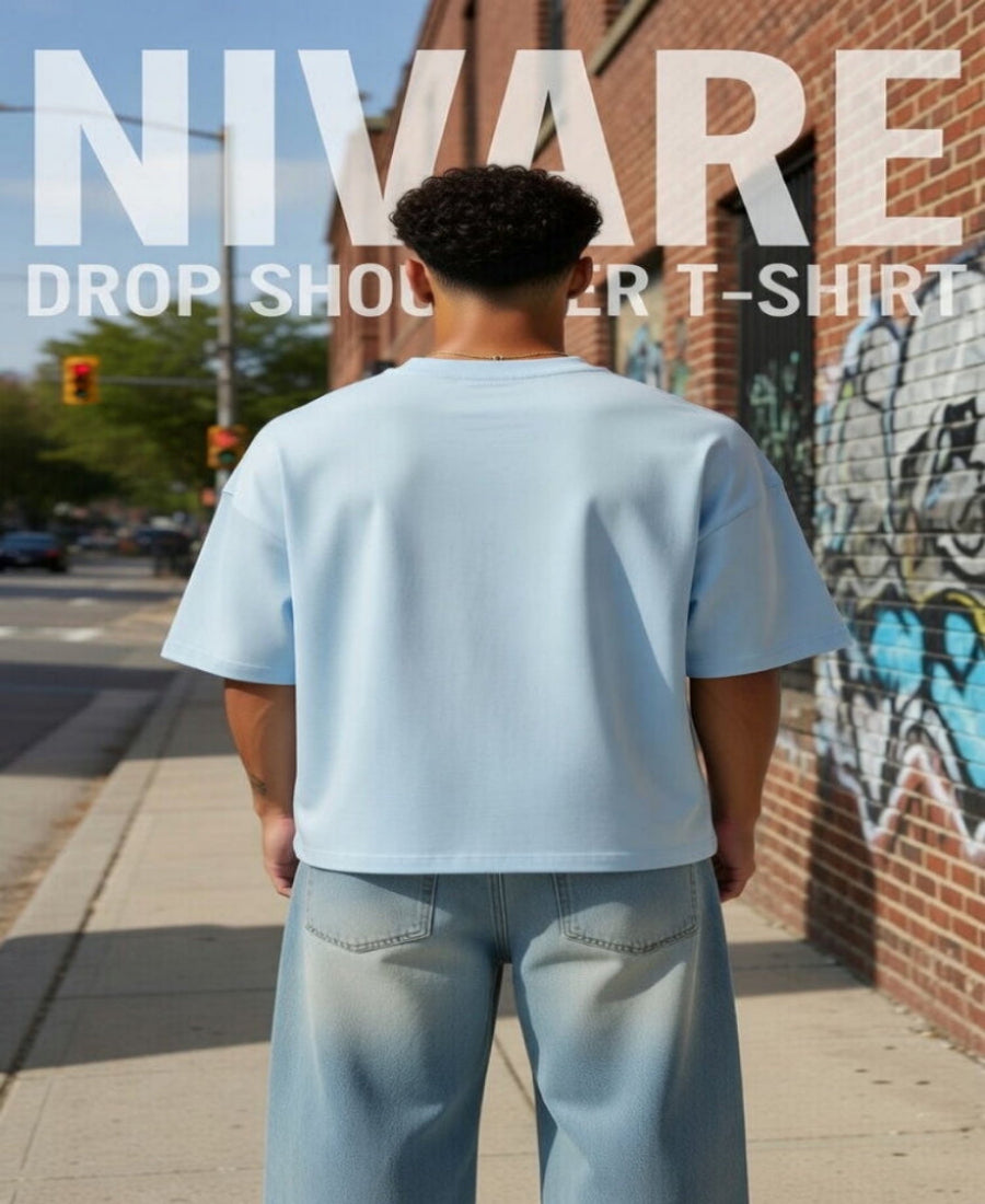 Person wearing a light blue t-shirt and jeans standing on a sidewalk with graffiti in the background, featuring 'NIVARE' branding.